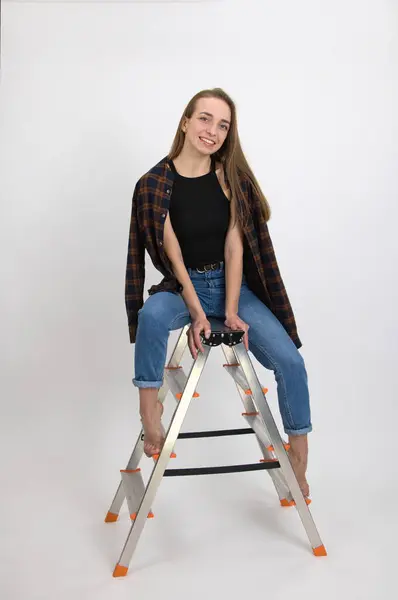 Beautiful blue-eyed smiling girl with long blond hair and bare feet expresses emotions of satisfaction while posing while sitting on a folding aluminum ladder. Close-up on a white background.