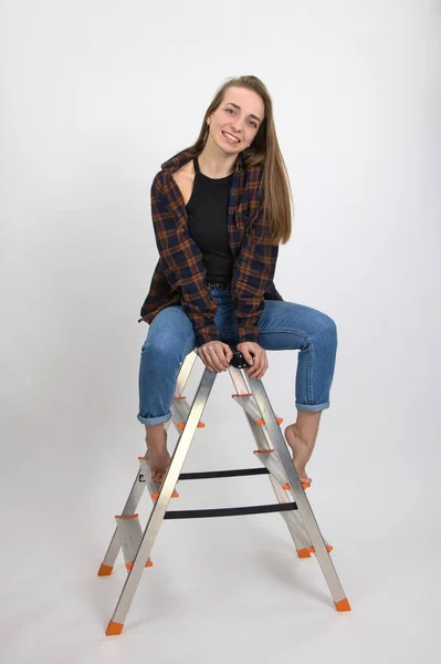 Beautiful blue-eyed smiling girl with long blond hair and bare feet expresses emotions of satisfaction while posing while sitting on a folding aluminum ladder. Close-up on a white background.