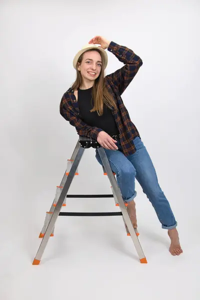 Beautiful barefoot blue-eyed smiling girl with long blond hair wearing a hat expresses emotions of satisfaction while posing while sitting on a folding aluminum ladder. Close-up on a white background.