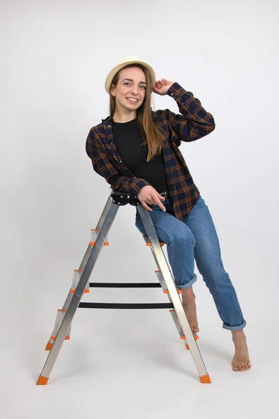 Beautiful barefoot blue-eyed smiling girl with long blond hair wearing a hat expresses emotions of satisfaction while posing while sitting on a folding aluminum ladder. Close-up on a white background.