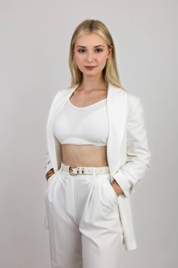 Uzun saçlı, beyaz takım elbiseli, elleri pantolonunun içinde olan sarışın kendinden emin bir görünüm sergiliyor. Beyaz bir arkaplanda izole edilmiş.