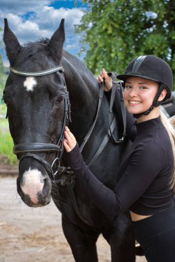 Çok güzel bir kız binici güzel gülüşlü ve sarı saçlı siyah bir atın yanında poz veriyor..