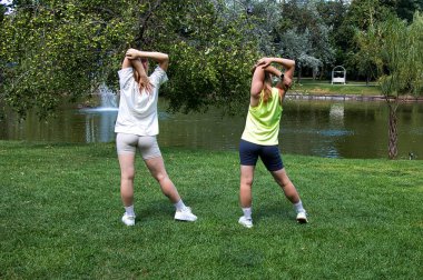 Spor giyimli iki genç kadın parkta esneme egzersizleri yapıyor. Yaz güneşli bir gün.