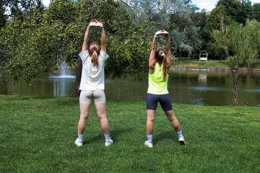 Spor giyimli iki genç kadın parkta esneme egzersizleri yapıyor. Yaz güneşli bir gün.