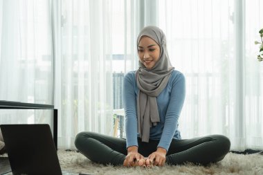 Asyalı genç kadın oturma odasında yoga yapıyor. Müslüman bir kadın laptopunda yoga videosuna bakıyor ve evde egzersiz yapıyor. Evde Fit Arap kadın eğitimi.