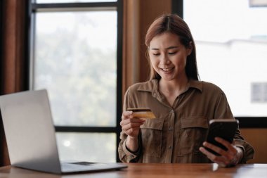 Genç Asyalı bir kadın internet üzerinden alışveriş yapıyor, cep telefonu ve kredi kartı kullanıyor, evden alışveriş yapıyor..