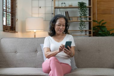 Gülümseyen yaşlı bayan akıllı telefonunu kullanıyor, internete bağlanıyor ve evdeki uygulamaları kullanıyor..