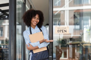 Kafe açmak için tabela çevirirken gülümseyen Afrikalı küçük işletme sahibi kadın. Mutlu Afro-Amerikalı garson önlüklü girişimci kapı açılışı tabelasında.