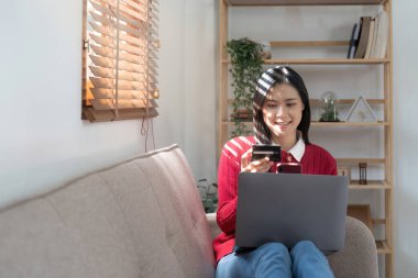 Oturma odasında dizüstü bilgisayar ve cep telefonu kullanırken kredi kartı tutan güzel bir genç kadın. Kara Cuma çevrimiçi alışveriş kavramı.