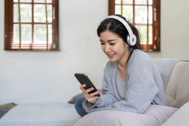 Asyalı kadın cep telefonu ve kulaklıkla müzik dinliyor. Sakin bir hafta sonu, kanepede otururken dinlenmek, favori melodi, popüler müzik, modern kablosuz kulaklık ve aletleri oturma odasında kullanmak..