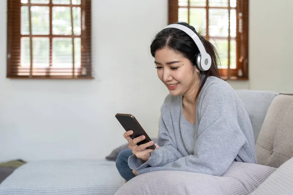 亚洲女人用手机和耳机听音乐 周末回家安静下来 坐在沙发上休息 欣赏最喜爱的曲调 流行音乐 在客厅里使用现代无线耳机和小玩意 — 图库照片