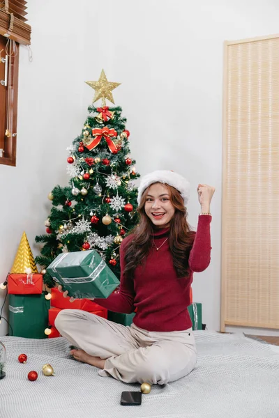 Happy woman sitting against Christmas tree background. Cheerful lady surprised of gift box. Marry Christmas and Happy Holidays.