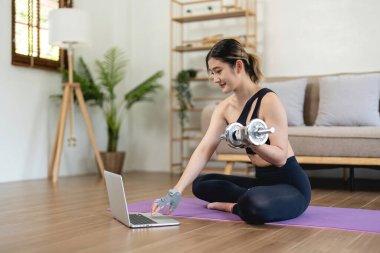 A sporty woman in sportswear is sitting on the floor with dumbbells using a laptop at home in the living room. Sport and recreation concept
