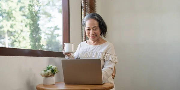 Happy aged Asian old woman working , sipping coffee while using laptop.