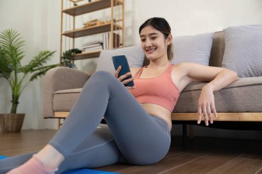 young woman exercising at home happily sitting and resting playing on the phone.