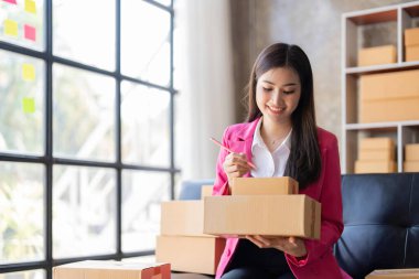 Young woman, an Asian online business owner, smiles, prepares parcel boxes and checks online orders for products to be delivered to customers.
