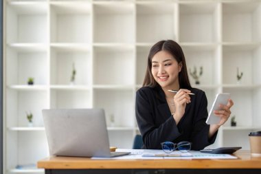 Asian businesswoman using smartphone to do accounting work with laptop and graph of financial reports in office.