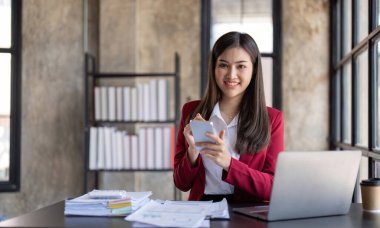 Asian businesswoman using smartphone to do accounting work with laptop and graph of financial reports in office.