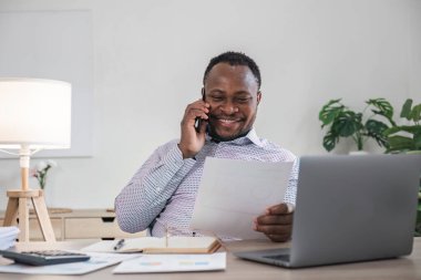 Başlangıç ofisinde cep telefonuyla konuşurken bilgisayarın başında oturan mutlu Afrikalı Amerikalı işadamı.