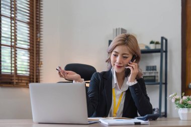 Mutlu genç Asyalı kadın cep telefonuyla konuşuyor ve ofisinde çalışırken gülümsüyor..