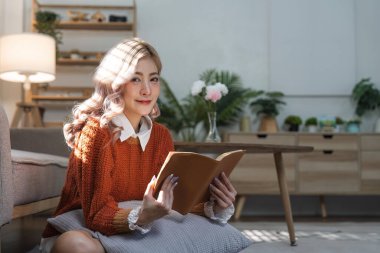 Genç Asyalı kadın rahat rahat kanepede oturup ilginç kitaplar okuyor. Oturma odasındaki kanepede mutlu kadın dinleniyor. Hafta sonları roman ya da hikayenin tadını çıkar..