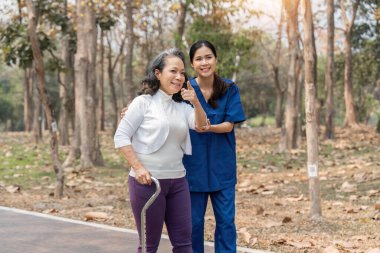 Hemşire yaşlı kadına Walker 'la yardım ediyor. Hemşire el ele tutuşuyor ve yaşlı bir kadının parkta yürümesine yardım ediyor..
