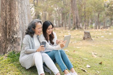 Güzel torun ve büyükanne yeşil parktaki ağacın altında oturup tabletten video izleyip, kahvelerini yudumluyorlar..