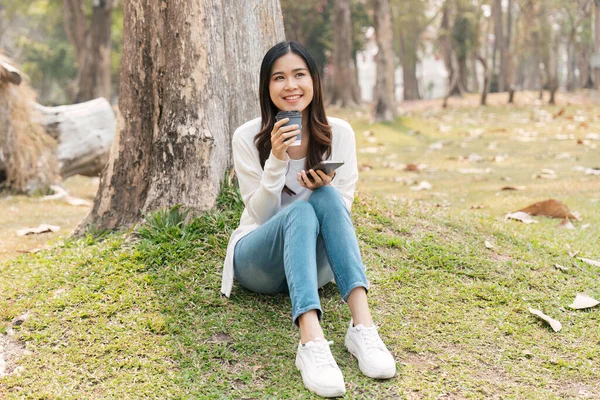 在户外公园里笑着的年轻女人休息一下 用手机上网看新闻 喝咖啡 — 图库照片