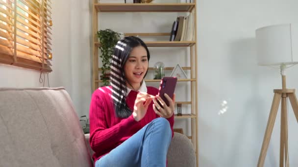 stock video Happy asian teen girl enjoy using mobile app sit on sofa, texting typing message hold smart phone look at cellphone screen browsing social media at home.