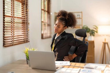 Ofiste dizüstü bilgisayarla çalışırken sırt ağrısı çeken genç bir afro kadın oturuyor..