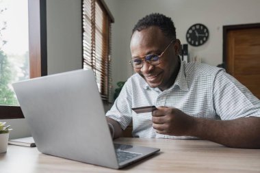 İnternetten kredi kartıyla ödeme yapan Afro-Amerikalı iş adamının internetten sipariş verirken görüntüsü. Başarılı siyah erkek, elinde plastik kartla dizüstü bilgisayar bankacılığı uygulaması kullanarak işlem yapıyor..
