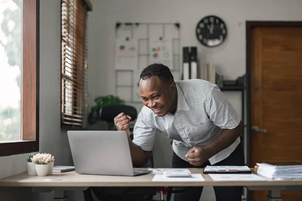Happy surprised African American businessman reading good news in letter, celebrating success, excited young man holding documents, sitting at work desk.