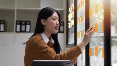 Genç Asyalı iş kadını cam duvardaki yapışkan kağıtları kullanarak proje planı üzerinde çalışıyor..