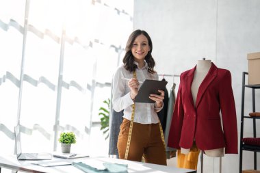 Gülümseyen kadın moda tasarımcısı atölyede dizüstü bilgisayar ve tabletle çalışıyor..