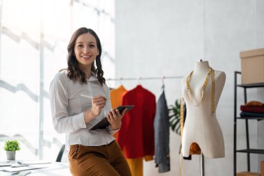 Gülümseyen kadın moda tasarımcısı atölyede dizüstü bilgisayar ve tabletle çalışıyor..