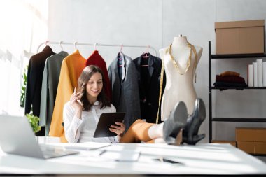 Gülümseyen kadın moda tasarımcısı atölyede dizüstü bilgisayar ve tabletle çalışıyor..