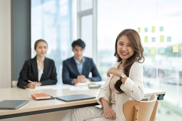 オフィスの腕の中に立つ若いアジア系ビジネス女性の肖像画が同僚との会話を背景に — ストック写真