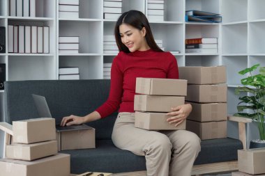 Kadın satıcı teslimat için paket kutusuna müşteri adreslerini yazar ve çevrimiçi mağazada, paket teslimatı kavramında ve çevrimiçi satış günlerinde dizüstü bilgisayarları kontrol eder..