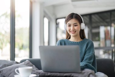 Modern kulaklıklı genç bir kadın oturma odasında kanepede oturur dizüstü bilgisayarla çalışır..