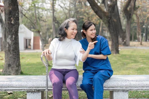 一位亚洲女性护理人员与一位老妇人坐在医院公园的长椅上聊天 — 图库照片