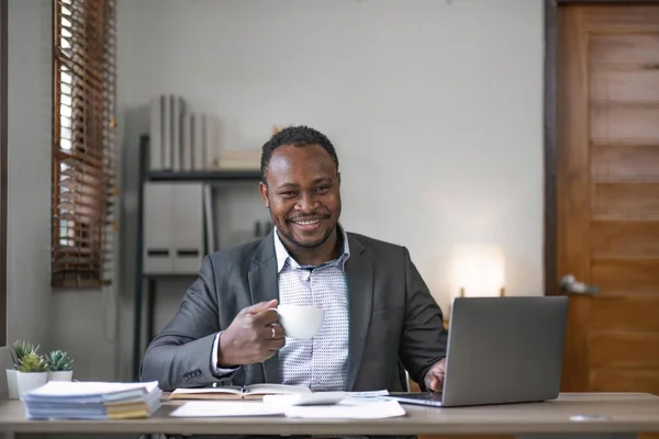 Young African black mansmiling businessman drinking coffee and using laptop in office, sitting at desk.