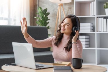 Evde dizüstü bilgisayar kullanan, kulaklıkla müzik dinleyen genç bir kadın. Kanepede oturan güzel, gülümseyen kız.