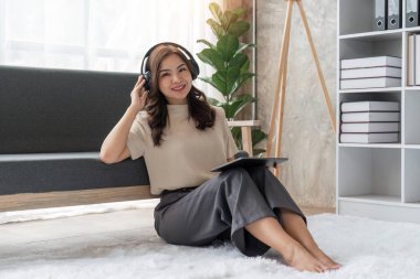 Evde tablet kullanan, kulaklıkla müzik dinleyen genç bir kadın. Kanepede oturan güzel, gülümseyen kız.