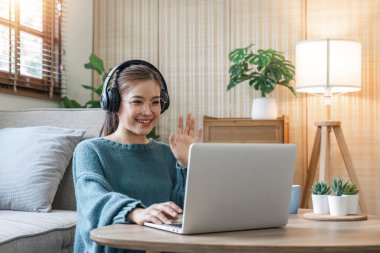 Kanepede oturmuş gündelik kıyafetlerle gülümseyen genç bir kadın bilgisayarla görüntülü konuşma, el sallama ev içinde dinlenme, insanların yaşam tarzı konsepti..