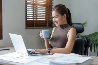 Güzel gülümseyen Asyalı kadın dizüstü bilgisayarda çalışıyor ve evdeki oturma odasında kahve içiyor. Asyalı iş kadını ev ofisinde finans ve hesap makinesinde çalışıyor..