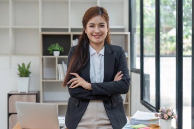 Young successful woman entrepreneur or an office worker stands with crossed arms near a desk in a modern office, looking at the camera and smiling.