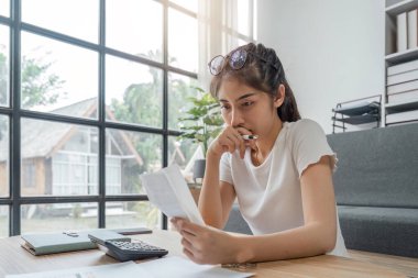 Evrakları elinde tutan, aile bütçesini hesaplayan, tasarruf yapmaya çalışan, stresli ve konsantre görünümlü genç bir kadın..