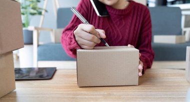 Asyalı bir kadın bir e-ticaret şirketi, onlara göndermeden önce müşterilerin bir listesini kağıda yazıyor. Web sitelerinde ve sosyal medyada bir e-ticaret şirketi işletiyor. Çevrimiçi ürün satma kavramı.