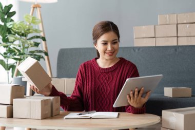 Paket kutuları tutan ve tabletten sipariş alan Asyalı bir kadın, bir online mağazası var, özel bir nakliye şirketi aracılığıyla paketleyip gönderiyor. Çevrimiçi satış ve online alışveriş kavramları.