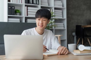 Genç adam dizüstü bilgisayarını kullanarak evde çalışıyor, online dersler alıyor ve notlar alıyor, gülümsüyor..
